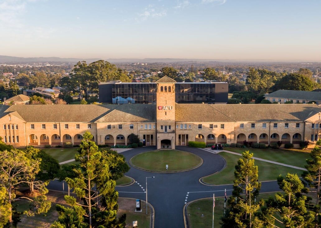 Aerial view of an ACU campus at golden hour, with a sandstone-brick building, arched colonnades and a central clock tower bearing the ACU logo, facing a circular driveway, lawns and pine trees.