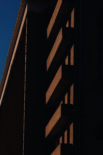Low-angle view of a dark modern building facade, warm light striping across its balconies against a deep blue sky